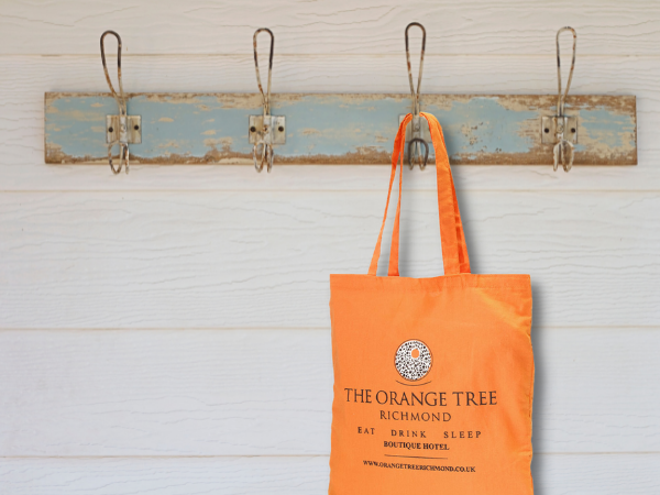 orange cotton tote bag, printed with The Orange Tree Richmond, handing on rustic hooks against white wall